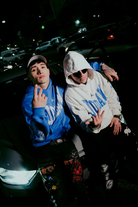 Dos personas con ropa azul y blanca junto a un automóvil de noche.