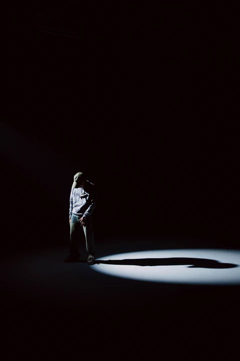 Figura de pie bajo un haz de luz, rodeada por oscuridad.