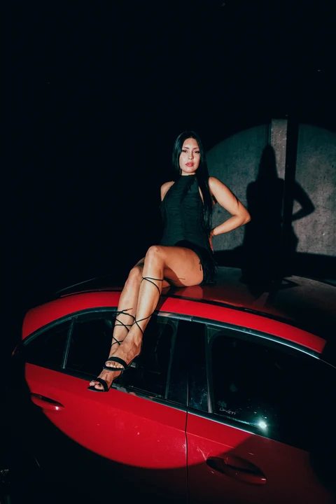 Mujer sentada sobre un automóvil rojo en una escena nocturna.
