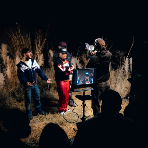 Equipo de rodaje junto a artistas y un monitor en un exterior nocturno.