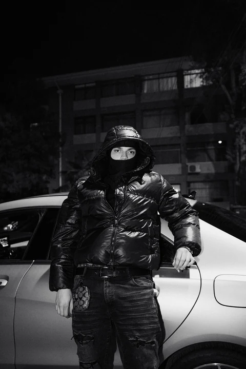 Figura con chaqueta y gorra negras frente a un automóvil, en blanco y negro.