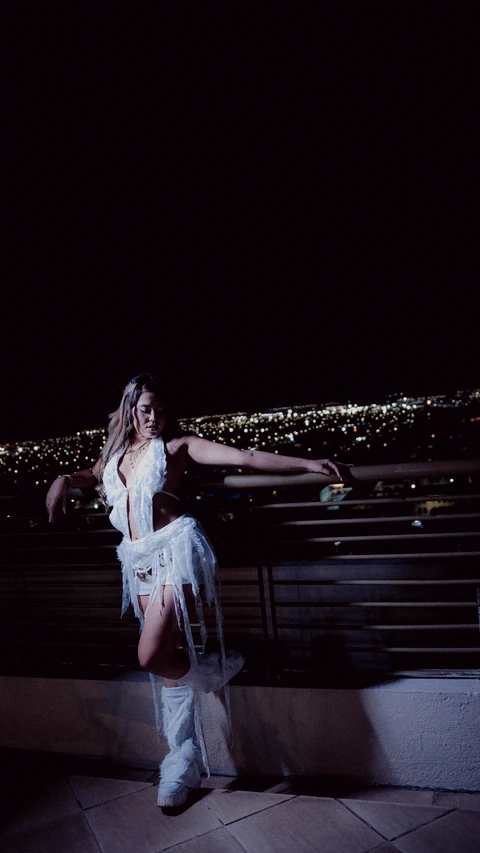 Mujer vestida de blanco junto a una baranda con luces de ciudad al fondo.