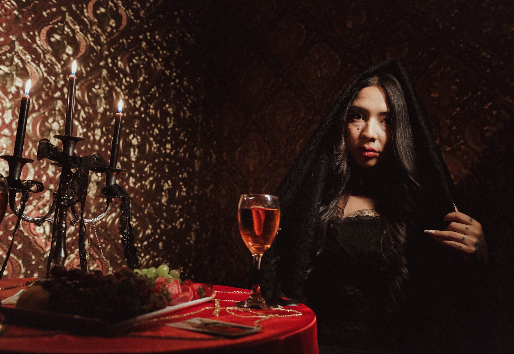 Mujer con velo negro y copa de vino en una mesa roja con candelabros, set del videoclip Olvídame tú de CloudSix